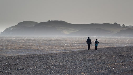 A silhouetted couple walking along a shingle beach towards a hill in the distance